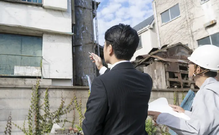 建物の前で調査中の背広姿の男性とヘルメット姿の作業員